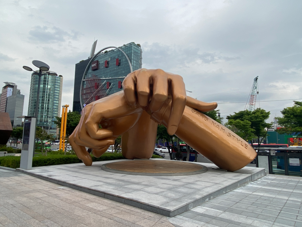 Large bronze statue of two crossed hands forming a dance move from the hit song “Gangnam Style,” located in a busy plaza in Seoul's Gangnam district. The base is engraved with "GANGNAM STYLE," highlighting a pop culture landmark in the area of where to stay in Seoul South Korea.