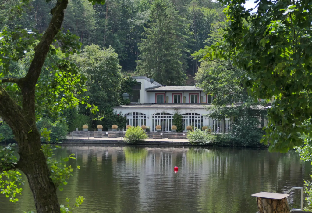A peaceful lakeside view of a white villa-style building surrounded by dense forest near Kaiserslautern. The calm water reflects the trees and structure, with a single red buoy floating in the foreground.