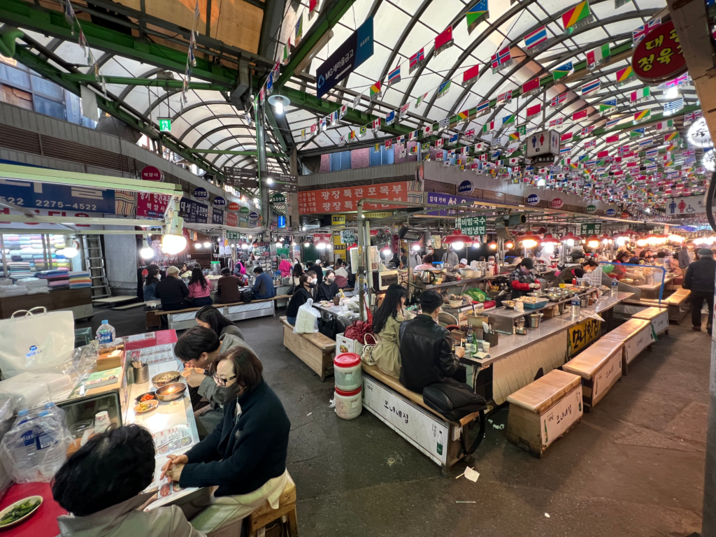 Indoor scene of Gwangjang Market in Seoul, where rows of food stalls are packed with people enjoying traditional Korean dishes under a ceiling decorated with international flags. Exploring this lively market is a flavorful and authentic thing to do in Seoul South Korea.