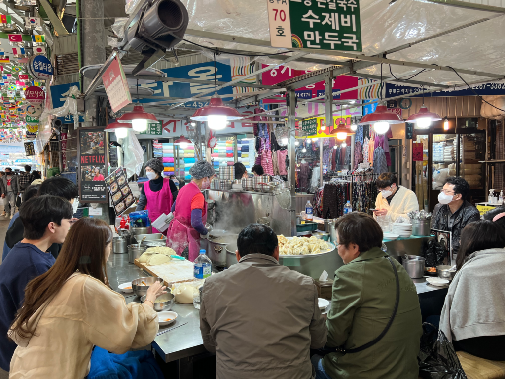 Close-up view of diners seated at a busy food stall in Gwangjang Market as vendors prepare hot dumplings and noodle soup in steaming pots. Sampling street food at this market is a delicious and popular thing to do in Seoul South Korea.