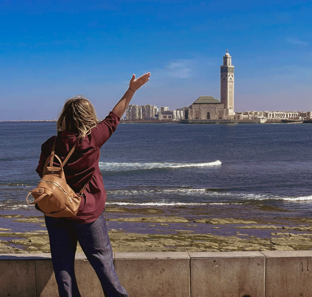 Woman with outstretched arms in a red shirt and linen pants with a tan bag with outstretched arms infront of the Hassan II Mosque in Casablanca