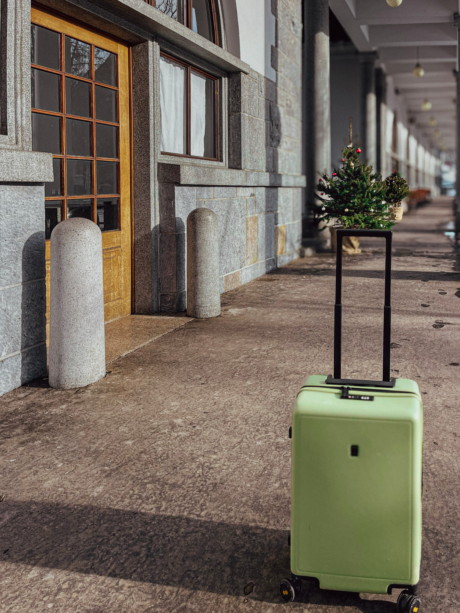 A green Level8 Luminous 20-inch carry-on suitcase stands upright on a stone walkway outside a building with arched wooden doors and stone walls. A small decorated Christmas tree on a stand adds a festive touch to the background.