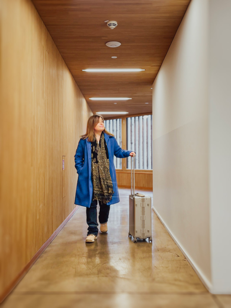 A woman in a blue coat walks through a modern hallway with wood-paneled walls, pulling a silver Level8 Gibraltar 20-inch carry-on suitcase. She looks upward as she moves forward, capturing a moment of travel or arrival indoors.