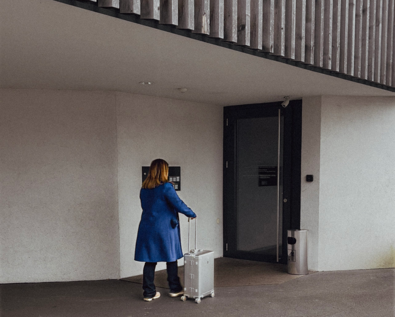 A woman wearing a blue coat stands outside a modern building, pulling a silver 20-inch Level8 Gibraltar carry-on suitcase. She faces a dark entry door with an intercom panel, suggesting she's about to check in or enter.
