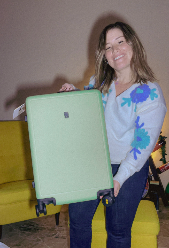 Smiling woman holding up a light green Level 8 Luminous Textured Carry-On 20", showcasing its smooth, minimalist design and black spinner wheels with yellow accents. The suitcase’s hard-shell exterior has a subtle matte texture for a stylish and durable finish.
