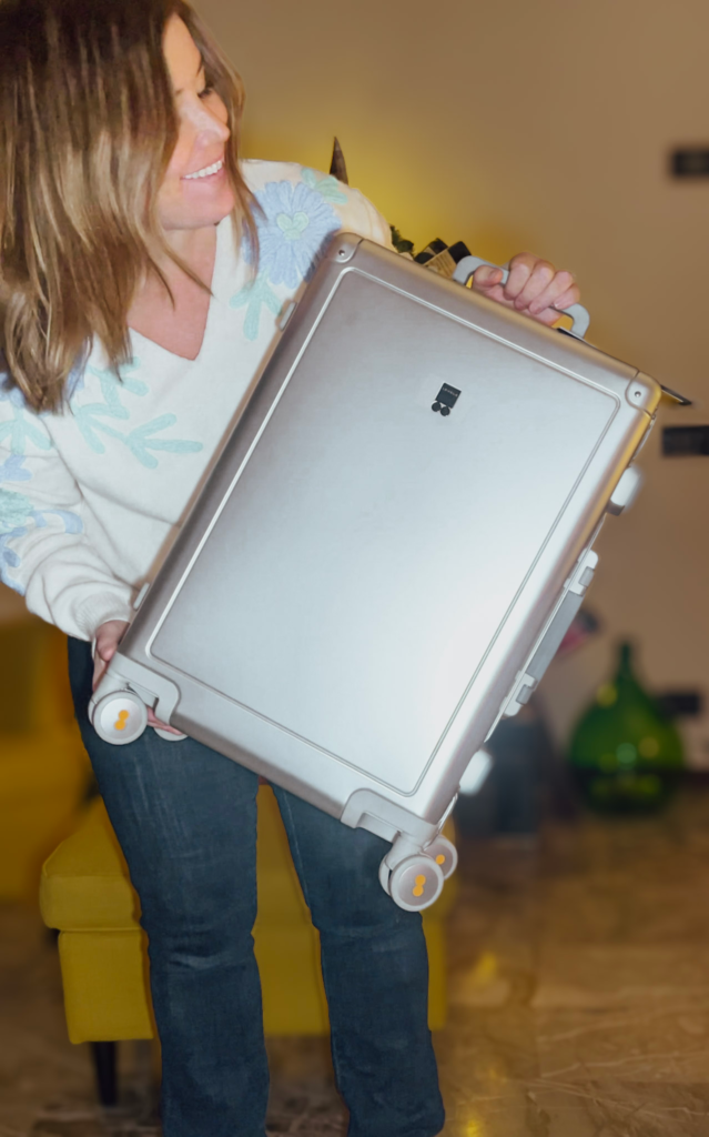 Smiling woman holding up a silver Level 8 Gibraltar Aluminum Carry-On 20" to show its smooth hard-shell exterior and spinner wheels with orange accents. The luggage appears lightweight and compact, ideal for easy handling.