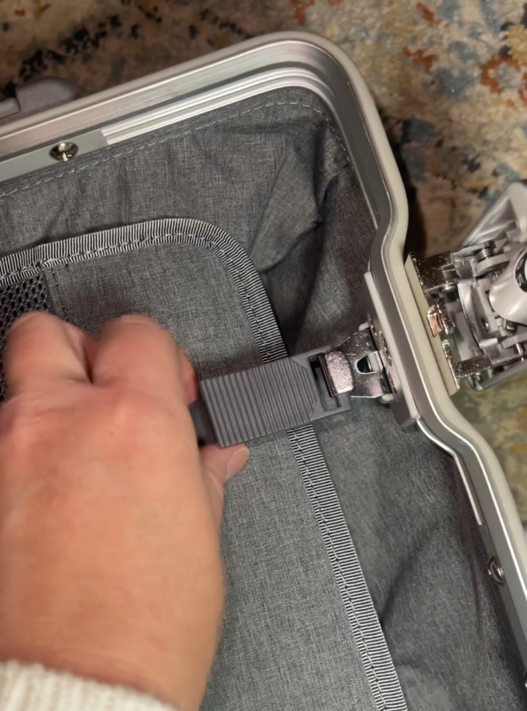 A hand clasps the interior strap buckle of the Level8 Gibraltar 20-inch carry-on, revealing its durable grey fabric lining and metallic frame construction.