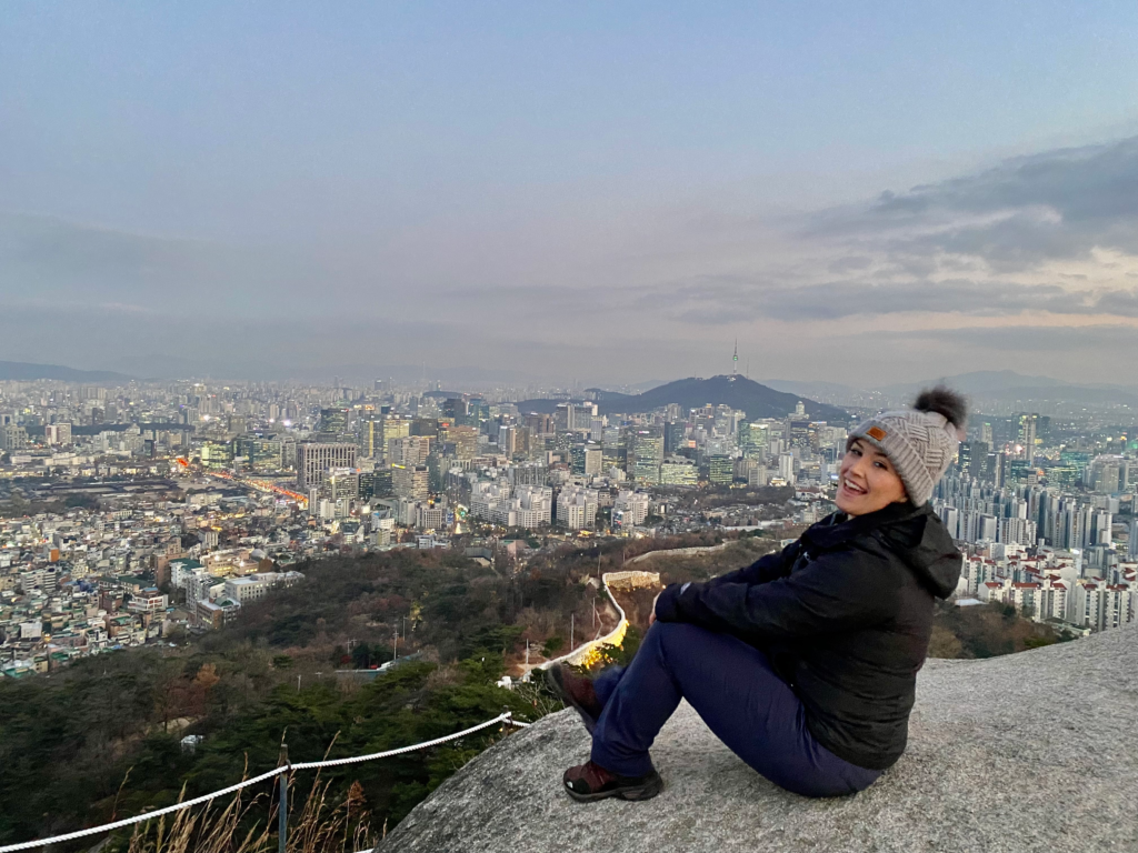 A smiling woman in winter gear sits on a rocky ledge of Inwangsan Mountain overlooking the expansive skyline of Seoul at dusk, with Namsan Tower visible in the distance. Scenic viewpoints like this are often accessible from nearby stays in Seoul South Korea that blend urban and natural landscapes.