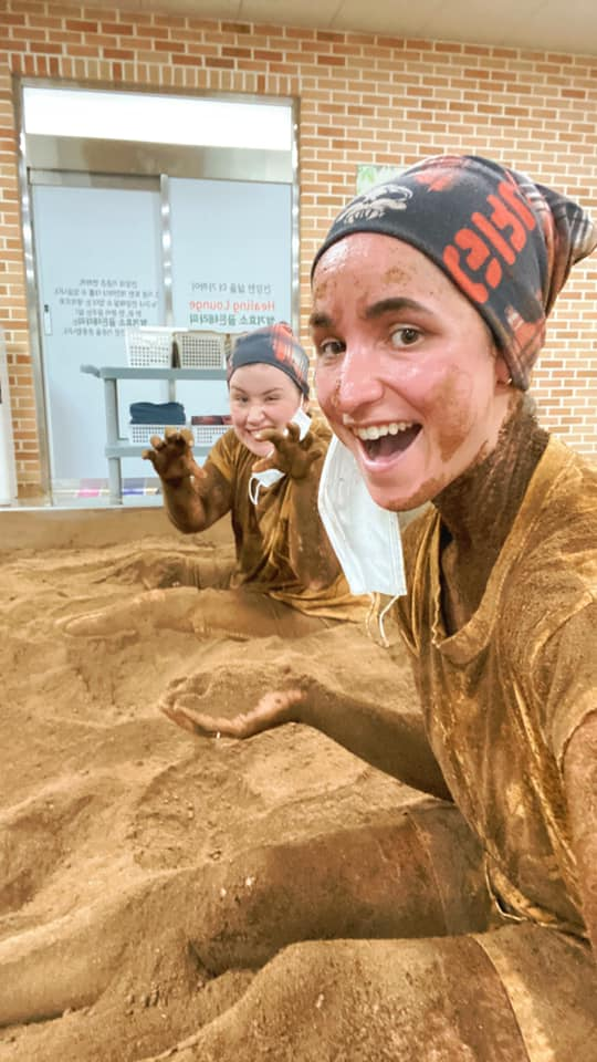 Close-up selfie of two women grinning and playfully showing their rice branl-covered hands while immersed in the brown soil bath at Chorok Miso Village. The background shows a spa area with brick walls and Korean signage.