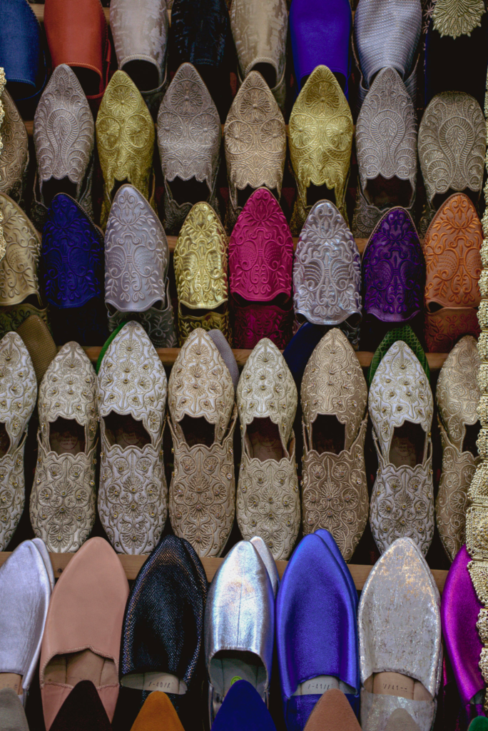 Rows of pointed Moroccan leather slippers displayed tightly together in a market stall in the Marrakech medina. The shoes feature intricate embroidery and rich colors including gold purple blue and cream showing the variety of traditional craftsmanship.