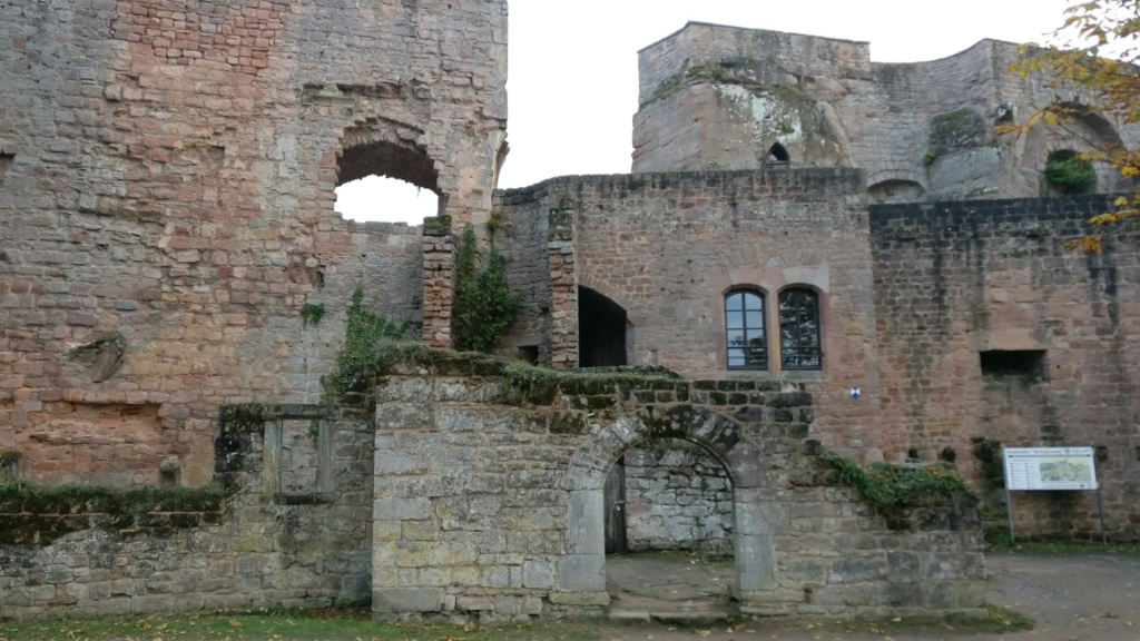 Close-up of weathered stone walls and archways at a historic castle ruin near Kaiserslautern. Ivy climbs parts of the old structure, with a signboard visible to the side indicating visitor information.