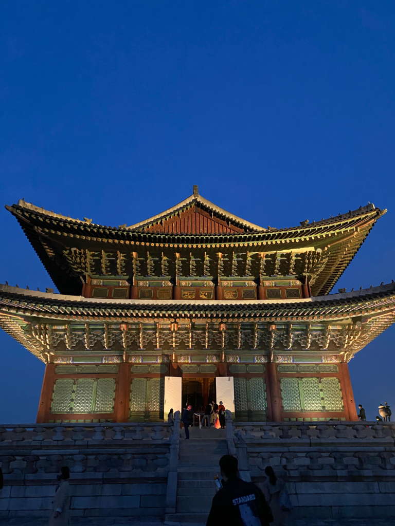 The main gate of Gyeongbokgung Palace glows under deep blue evening skies, with visitors climbing the steps toward the grand wooden structure. This historic site is near several traditional and modern accommodation options for those exploring where to stay in Seoul South Korea.