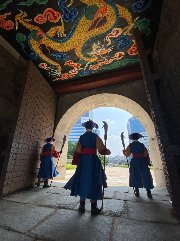 Three traditional Korean guards in blue and red uniforms stand under a stone archway at a historic gate in Seoul, with a vibrant dragon painting on the ceiling above them. The view opens to a modern cityscape, blending history with the urban environment of Seoul South Korea.
