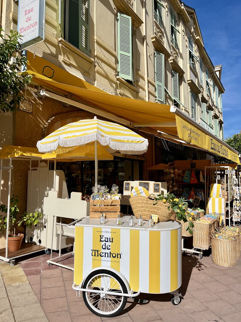 Bright yellow Eau de Menton cart outside boutique in sunny French street.