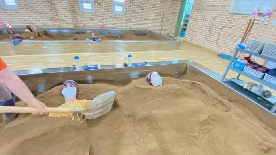 Two people lie mostly buried in a bed of warm soil with only their heads visible, wearing face masks and head wraps, while a staff member uses a shovel to cover them further at Chorok Miso Village. The setting has multiple soil beds and a clean, spa-like atmosphere with brick walls.