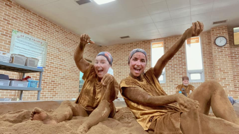 Two joyful women pose with raised arms while sitting in communal rice bran pit, their clothes and skin coated in brown soil at Chorok Miso Village. A third person sits in the background, reinforcing the group wellness experience.