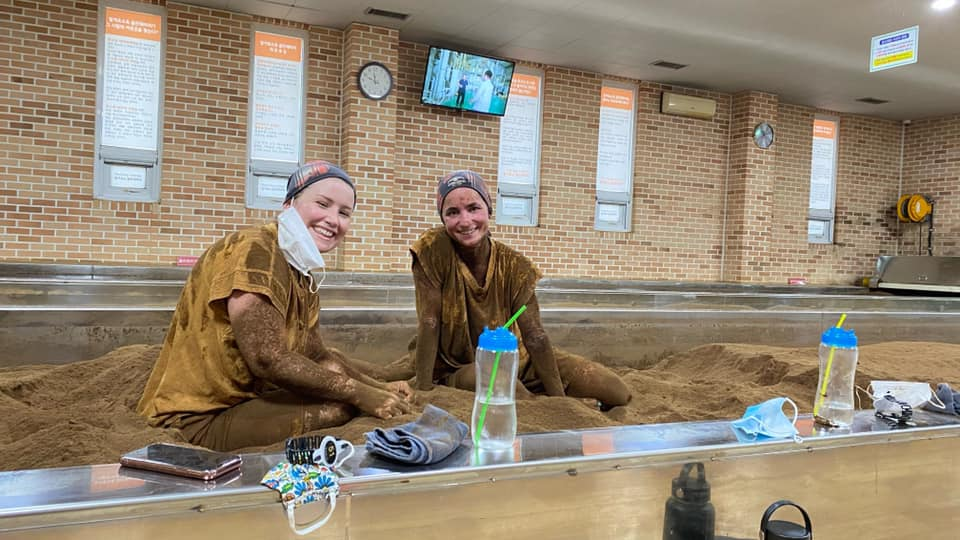 Two women, covered in rice bran and smiling, sit up from a communal rice bran bed inside Chorok Miso Village's wellness facility. They wear matching brown outfits and head wraps, with water bottles and personal items lined up on the edge of the pit.