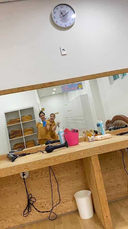 Mirror selfie in the locker room at Chorok Miso Village shows two women in matching brown spa uniforms smiling and holding water bottles. The counter is stocked with hair dryers, toiletries, and personal care items.