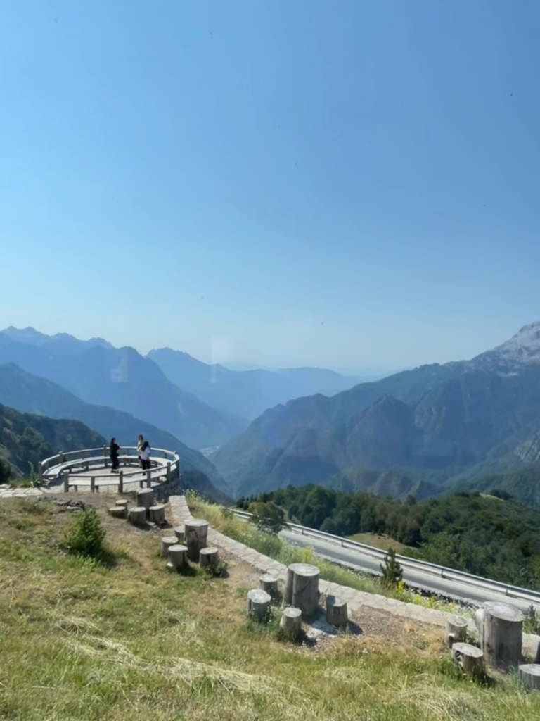 Circular wooden viewing platform overlooking a vast mountain valley with layered blue peaks in the distance. Two people stand at the railing taking in the dramatic Albanian Alps landscape.