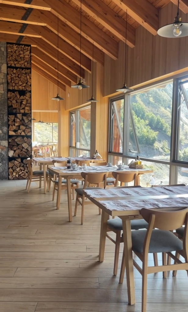 Warm wooden dining room with exposed beams, pendant lights, and large windows overlooking mountain scenery. Simple wooden tables are set for breakfast, creating a cozy alpine guesthouse atmosphere.