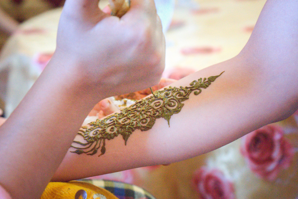 A close-up of a womans arm being decorated with a detailed henna pattern in dark green paste, featuring swirls and floral elements.