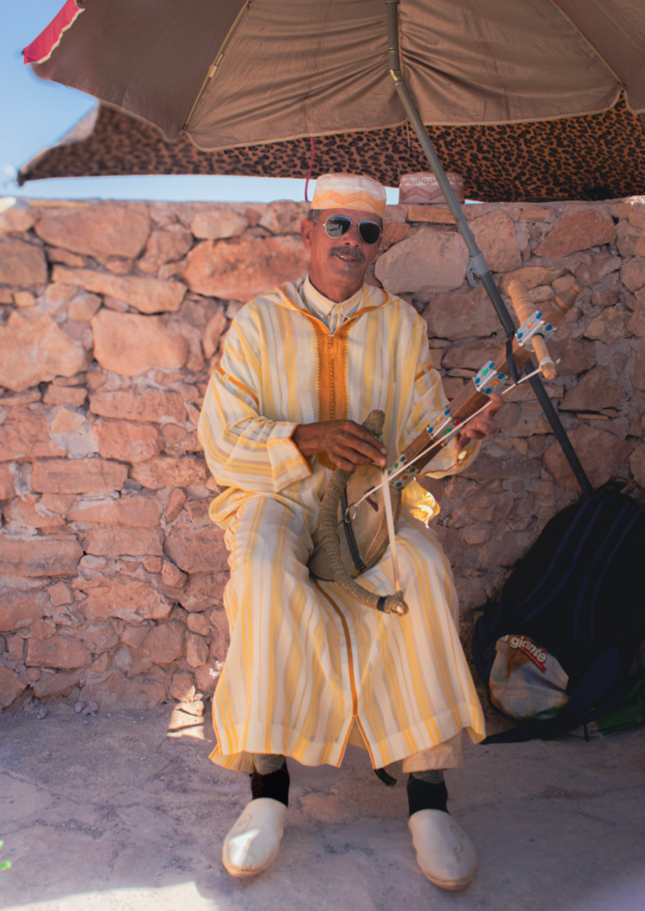 A man in traditional Moroccan clothing sits against a stone wall under a large umbrella in Ait Ben Haddou, holding a wooden string instrument decorated with colorful details. He wears a striped djellaba, sunglasses, and white slippers, capturing a relaxed street scene at the historic ksar.