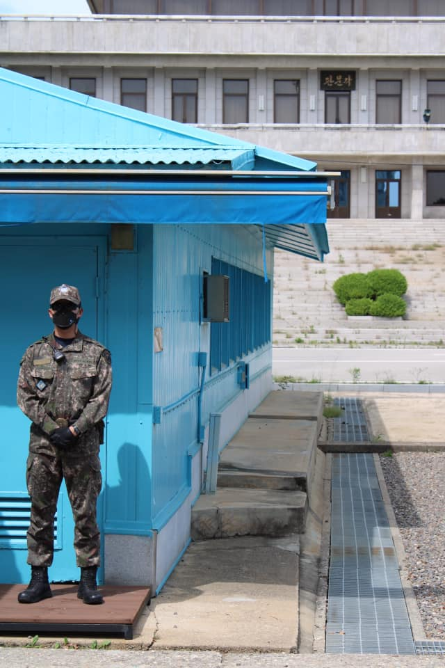 South Korean soldier standing at attention outside a bright blue building at the Joint Security Area with a government building visible behind him.