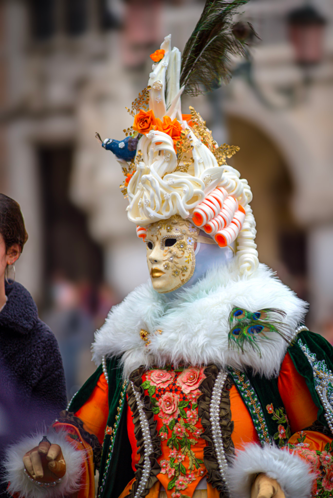 Person wearing an ornate Venetian carnival costume with a gold embellished mask and towering white curled wig decorated with orange roses and a peacock feather. The outfit features a white fur collar, green velvet cape, pearl strands, and a bright orange floral embroidered bodice, standing in a historic city square during a festival.