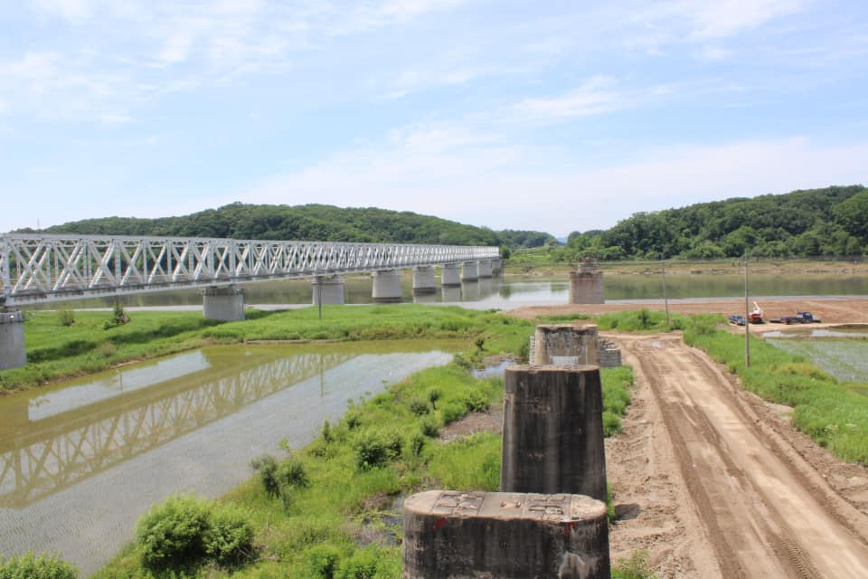 Wide view of a steel bridge spanning the river with old concrete supports in the foreground and green hills lining the horizon near the DMZ.