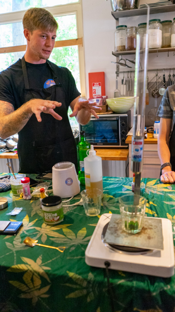 An instructor wearing a black apron gestures while explaining the infusion process at a table filled with bottles, a scale, and mixing tools. The scene shows the interactive and educational atmosphere of the Get Baked Amsterdam workshop in Amsterdam Netherlands.