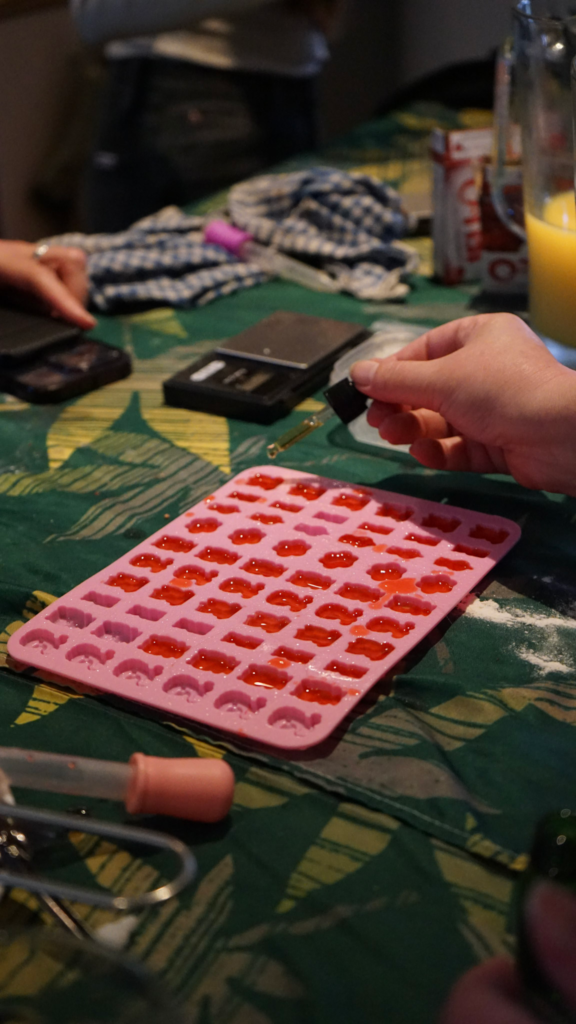 A hand uses a dropper to fill pink silicone molds with red liquid mixture while baking tools and ingredients sit on a patterned table. The image shows participants preparing infused treats during the Get Baked Amsterdam cannabis workshop.