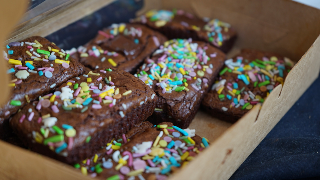 Close up of freshly baked brownies covered in rainbow sprinkles arranged inside a cardboard box. The image captures the final edible creations from the Get Baked Amsterdam cannabis workshop.