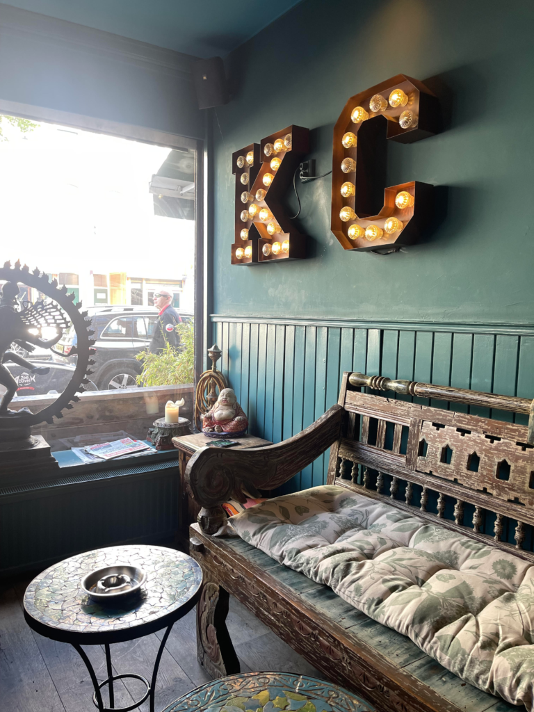 A cozy lounge area with a wooden bench, mosaic table, and illuminated KC wall letters inside a cannabis friendly space in Amsterdam. Natural light streams through the window, creating a relaxed atmosphere connected to the workshop experience.
