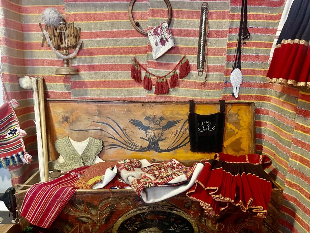 Colorful display of traditional Albanian women's clothing and accessories at the Women's Museum in Tirana, featuring embroidered vests, woven fabrics, and a painted chest with an eagle motif. A striped textile backdrop holds vintage tools, fringe, and decorative items showcasing local heritage.