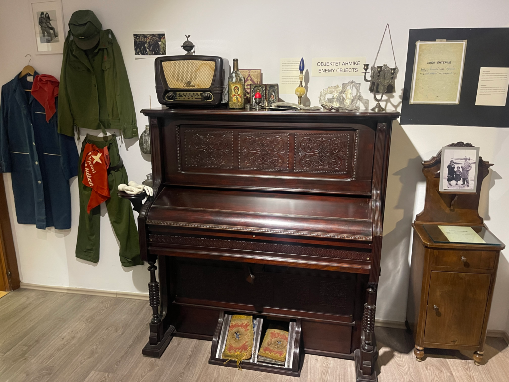 Exhibit at the Women’s Museum in Tirana showing a wooden upright piano surrounded by objects labeled enemy objects during the communist era. Military uniforms, a vintage radio, religious icons, and confiscated books are displayed to illustrate how personal belongings were seized under the regime.