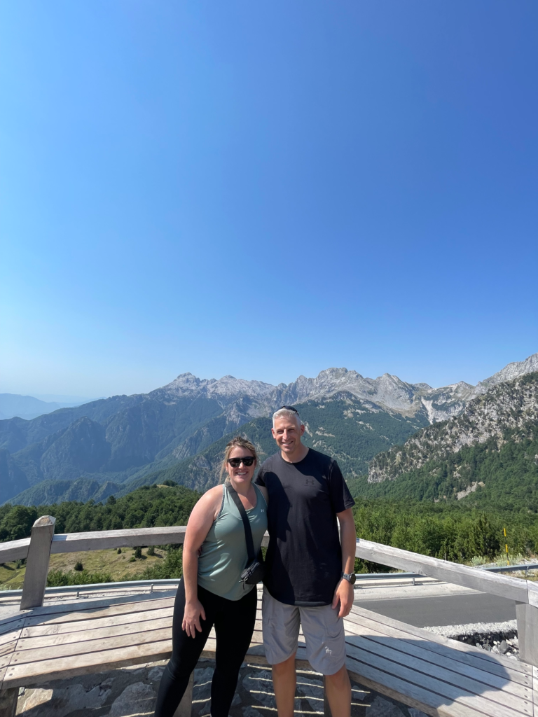 Couple standing on a wooden overlook with rugged mountains rising behind them under a clear blue sky. The dramatic peaks and deep green valleys stretch across the background.