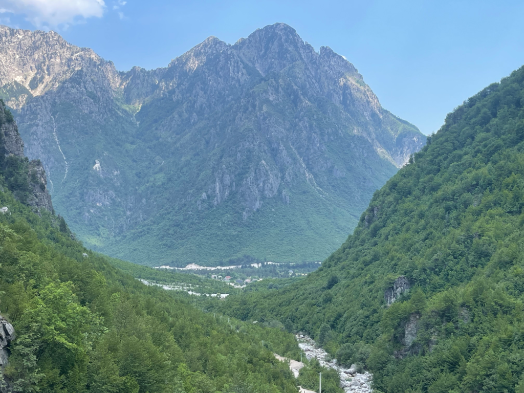 Expansive view over a lush green valley in Theth surrounded by steep rocky mountains. A small village and winding river are visible far below between the forested slopes.