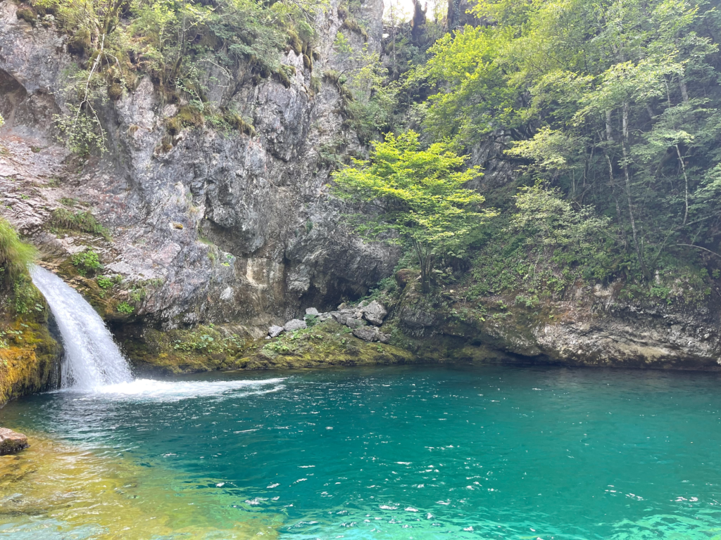 Small waterfall flowing into a the Blue Eye surrounded by rocky cliffs and dense green forest in Theth. Moss lines the rocks at the waters edge and the water appears calm and inviting beneath the trees.