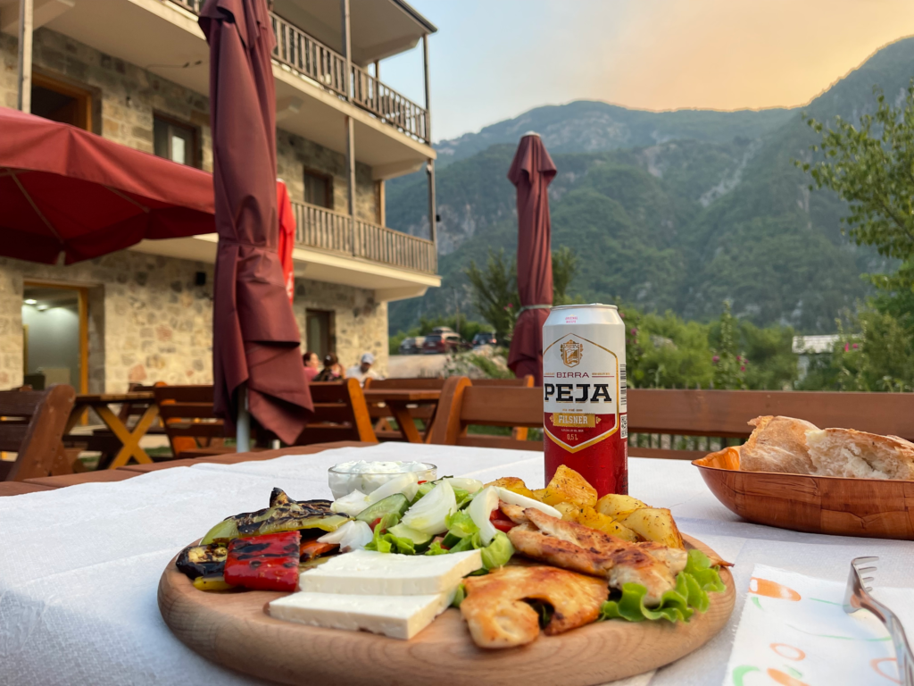 Plate of grilled chicken, roasted potatoes, fresh salad, grilled vegetables, and slices of white cheese served on a wooden board at a guesthouse in Theth. A can of Birra Peja sits on the table beside the meal, with mountain views and a stone lodge in the background at sunset.