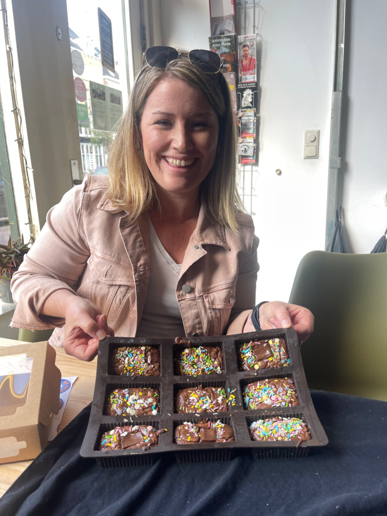 A smiling woman holds a tray of freshly made brownies topped with sprinkles while seated indoors. The image shows a participant proudly presenting her creations from the Get Baked Amsterdam cannabis workshop in Amsterdam Netherlands.