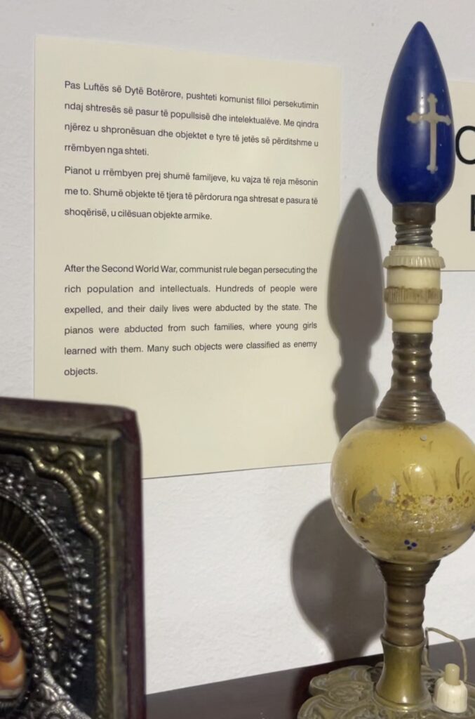 Museum display showing a vintage oil lamp with a blue glass top marked by a white cross beside a wall panel explaining post World War Two communist persecution in Albania. The text describes how wealthy families were expelled and personal belongings such as pianos were confiscated and labeled enemy objects.