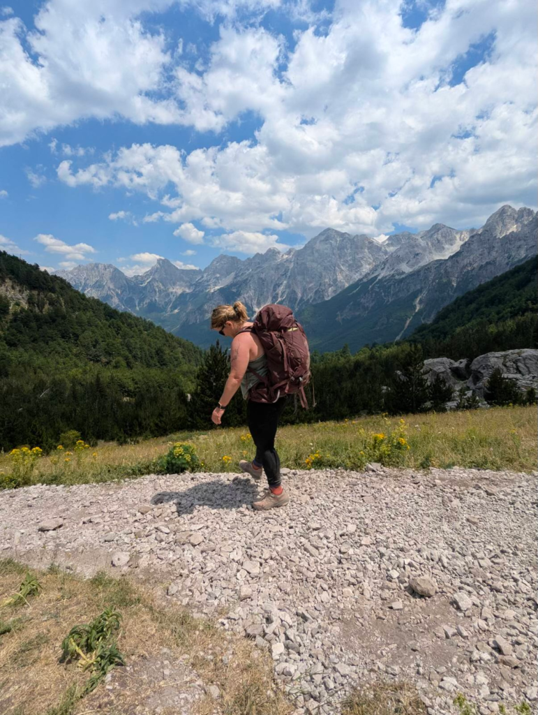 Hiker walking along a wide gravel path with a large backpack, framed by towering gray mountains and forested slopes. The open valley and layered peaks stretch across the horizon under scattered clouds.