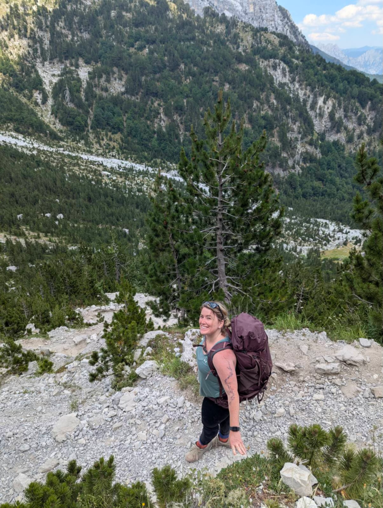 Smiling hiker with a large backpack standing on a rocky mountain trail surrounded by pine trees and steep valley walls. The rugged slopes of Valbone Valley rise behind her.