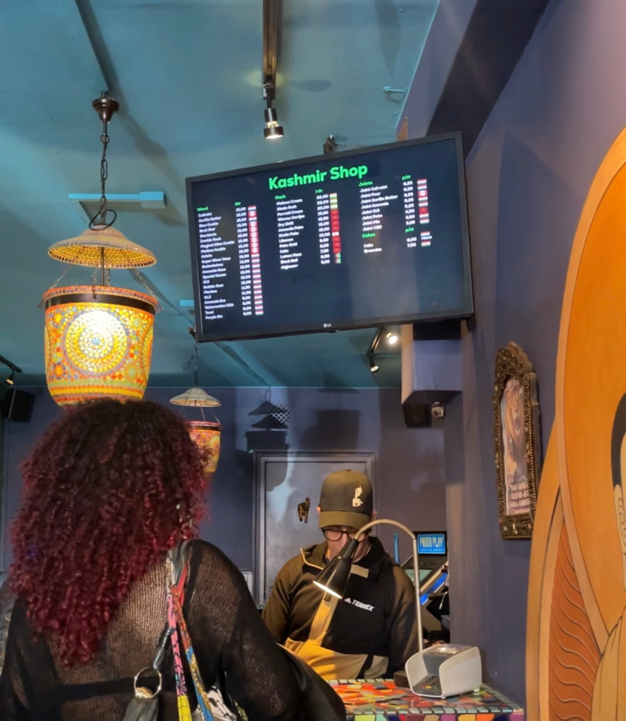 Inside Kashmir Shop in Amsterdam with a digital menu board overhead listing cannabis strains and prices. A customer stands at the counter beneath colorful hanging lamps in the cozy shop setting.