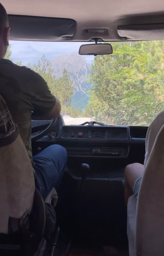Driver navigating a rugged dirt road through rocky scree fields toward a guesthouse in Valbone Valley after the Theth to Valbone hike in the Albanian Alps. View from inside the vehicle shows the dashboard and front seats framing pine trees and steep gray mountain peaks ahead, capturing the remote terrain and rough transfer conditions hikers experience after descending the trail.