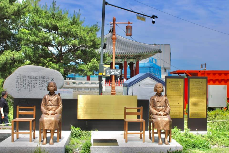 Statue memorial of two seated girls representing comfort women with empty chairs beside them and informational plaques in the background.