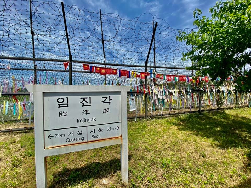 Sign reading Imjingak with distances to Gaeseong 22 kilometers and Seoul 53 kilometers standing in front of a barbed wire fence covered in colorful peace ribbons.