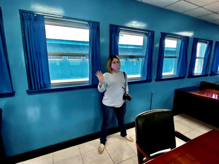 Woman wearing a pink face mask stands inside a bright blue conference room at the Joint Security Area raising her hand near a row of windows overlooking the DMZ.