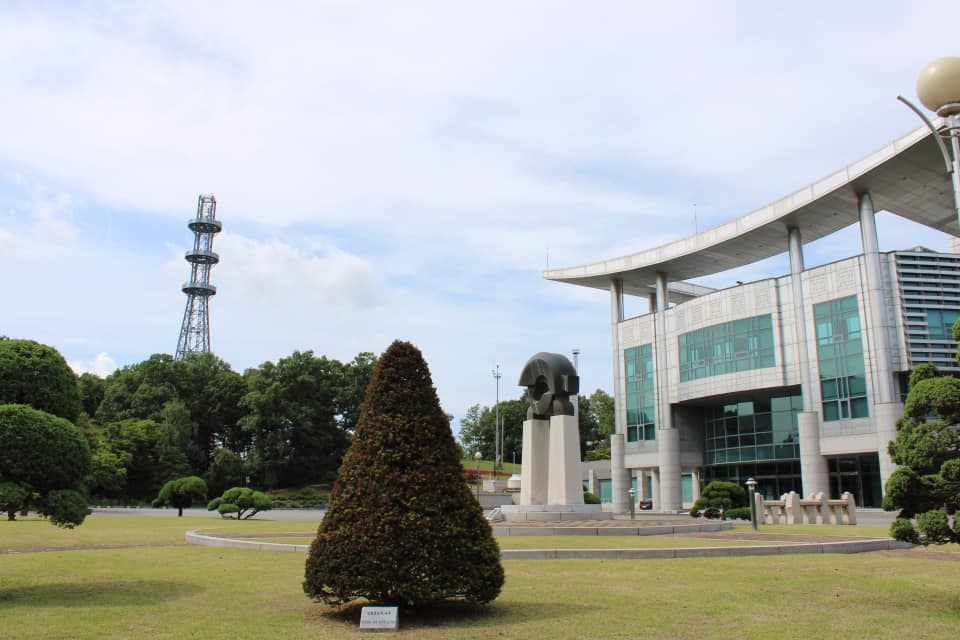 Modern building and sculpture on landscaped grounds near the Joint Security Area with a tall observation tower visible in the distance.