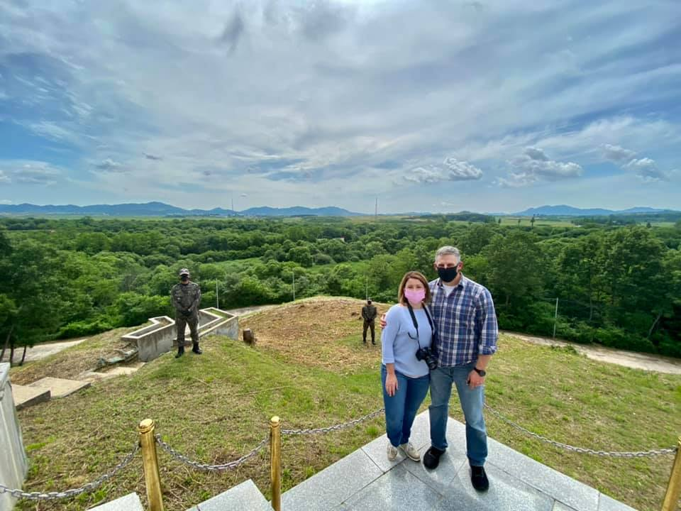Couple wearing face masks standing at an observation point overlooking the green landscape of the DMZ with soldiers spaced out behind them.